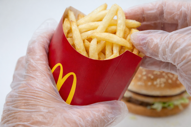 Gloved hands holding a McDonald’s French fries carton with a hamburger in the background, illustrating fast food packaging, plastic food handling, and potential phthalate chemical exposure risks linked to processed and packaged foods.