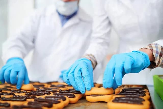 Workers wearing blue nitrile gloves inspect and handle chocolate-coated pastries on a production line, illustrating industrial food processing, packaging hygiene, and potential chemical exposure risks in commercial baked goods.